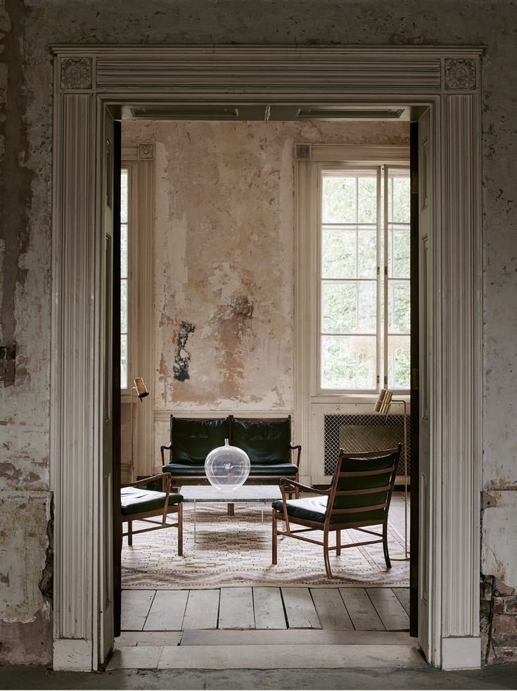 An enfilade through a panelled doorway into a room with stripped plaster, sash windows and a few pieces of furniture